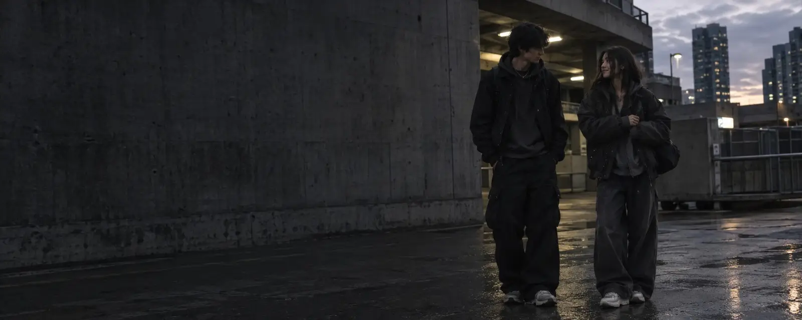 Two friends in oversized black streetwear walking across a wet concrete plaza at dusk